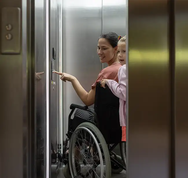 Frau im Rollstuhl fährt mit einem kleinen Mädchen im Aufzug und drückt einen Knopf.