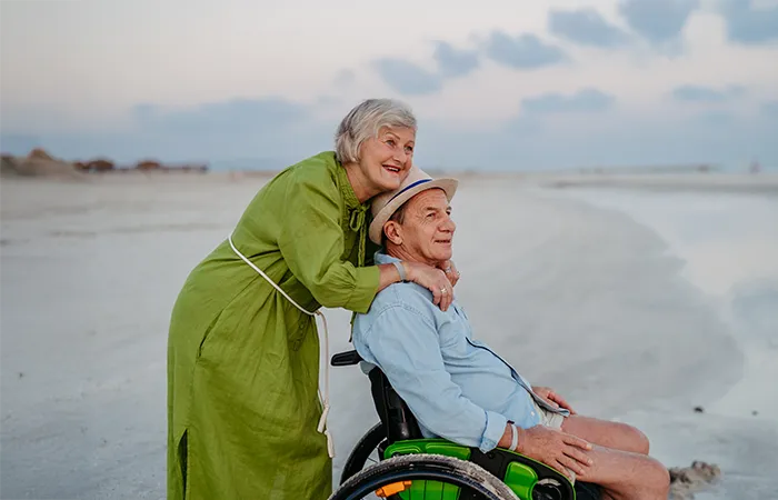Älteres Paar am Strand, Frau umarmt Mann im Rollstuhl, beide blicken lächelnd aufs Meer.