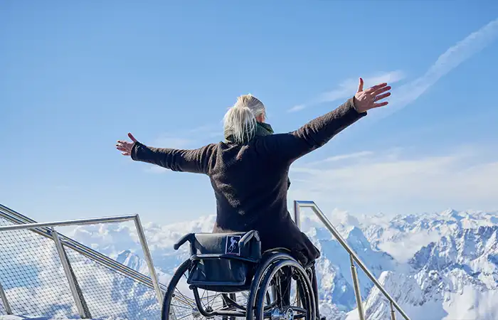 Eine Person im Rollstuhl streckt ihre Arme aus, während sie sich auf dem Gipfel der Zugspitze in Garmisch-Partenkirchen befindet. Im Hintergrund sind schneebedeckte Berge und ein strahlend blauer Himmel zu sehen.