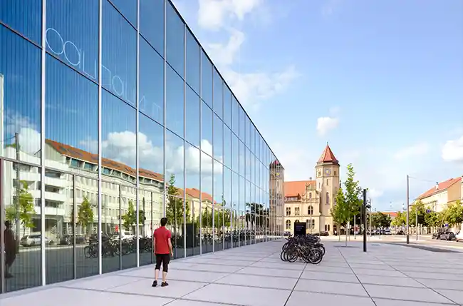 Das Bauhaus-Museum Dessau hat eine große Glasfassade, in der sich die umliegenden Gebäude spiegeln. Eine Person geht auf das Museum zu.