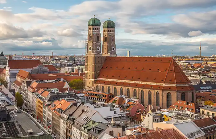 Frauenkirche, eine große Kirche in München, mit zwei hohen Türmen, die von grünen Kuppeln gekrönt werden. Die Kirche ist von vielen Gebäuden mit roten und braunen Dächern umgeben.
