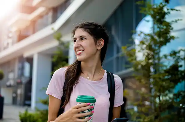 Junge Frau mit Hörgerät hält Kaffee und Smartphone in der Hand.