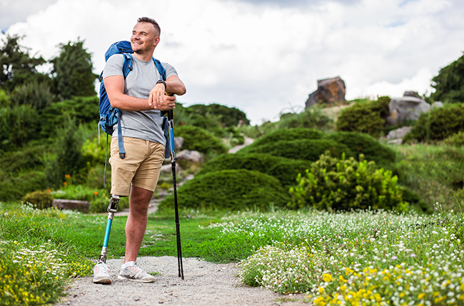 Mann mit Beinprothese wandert mit Rucksack und Stöcken durch grüne Landschaft, lächelnd im Sonnenschein.