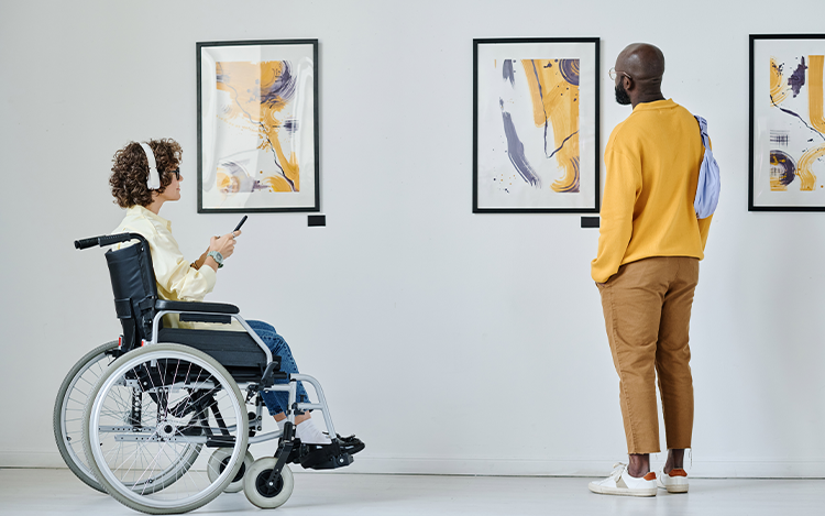 Person im Rollstuhl betrachtet mit Begleitung abstrakte Gemälde in heller Galerie.