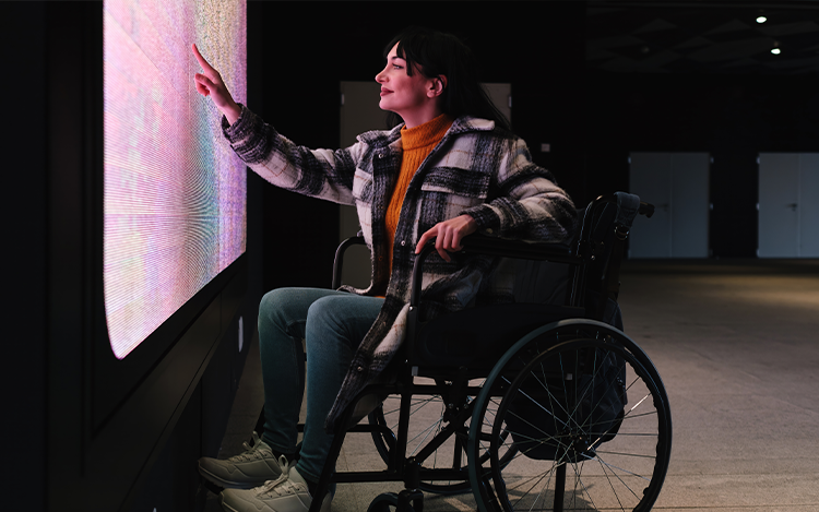 Frau im Rollstuhl berührt interaktive, leuchtende Bildschirmwand in dunklem Ausstellungsraum