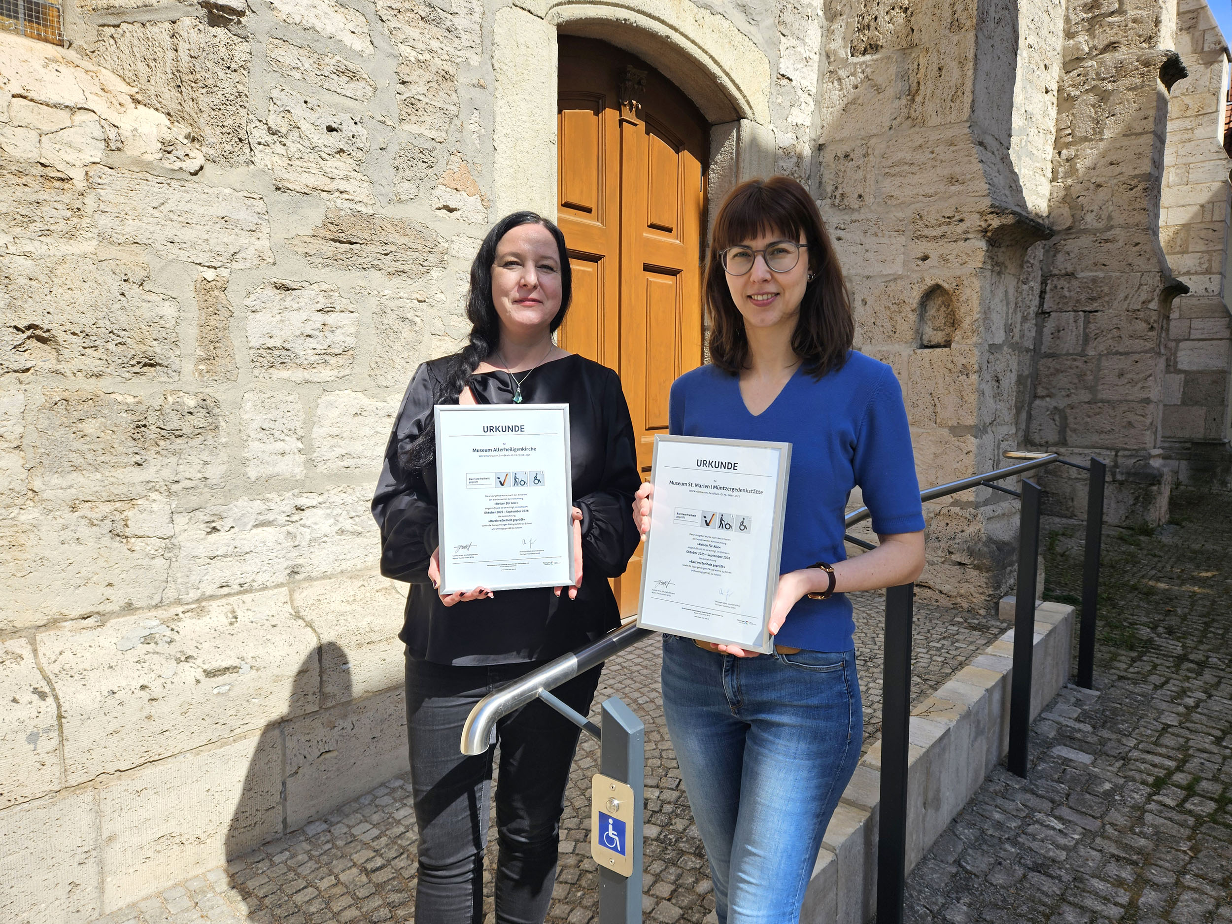 Zwei Frauen stehen vor einer Wand und halten beide eine "Reisen für Alle"-Urkunde hoch