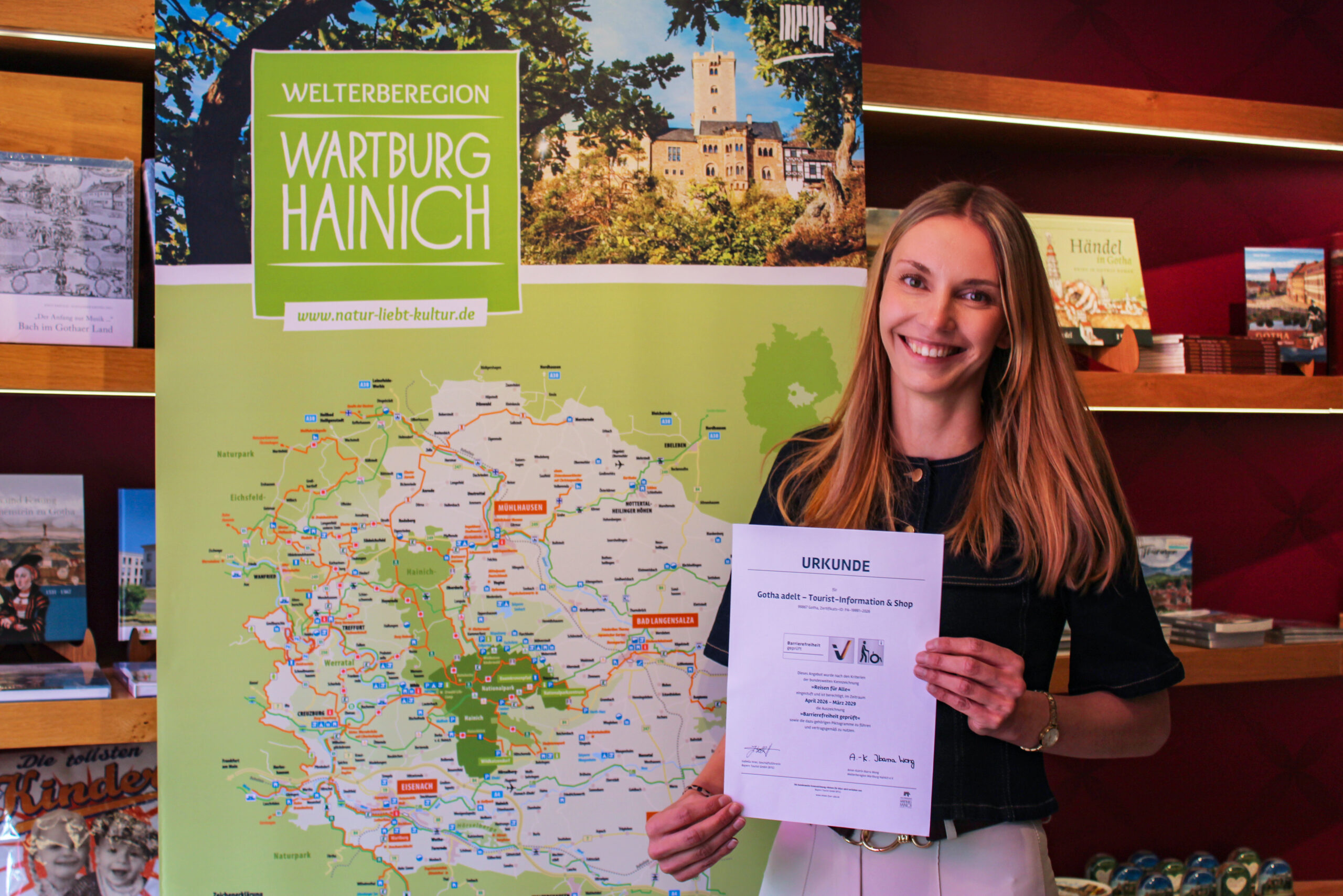 Eine Frau hält die Urkunde „Reisen für Alle“ der Tourist-Information Gotha vor einer Karte der Welterberegion Wartburg Hainich in die Kamera.