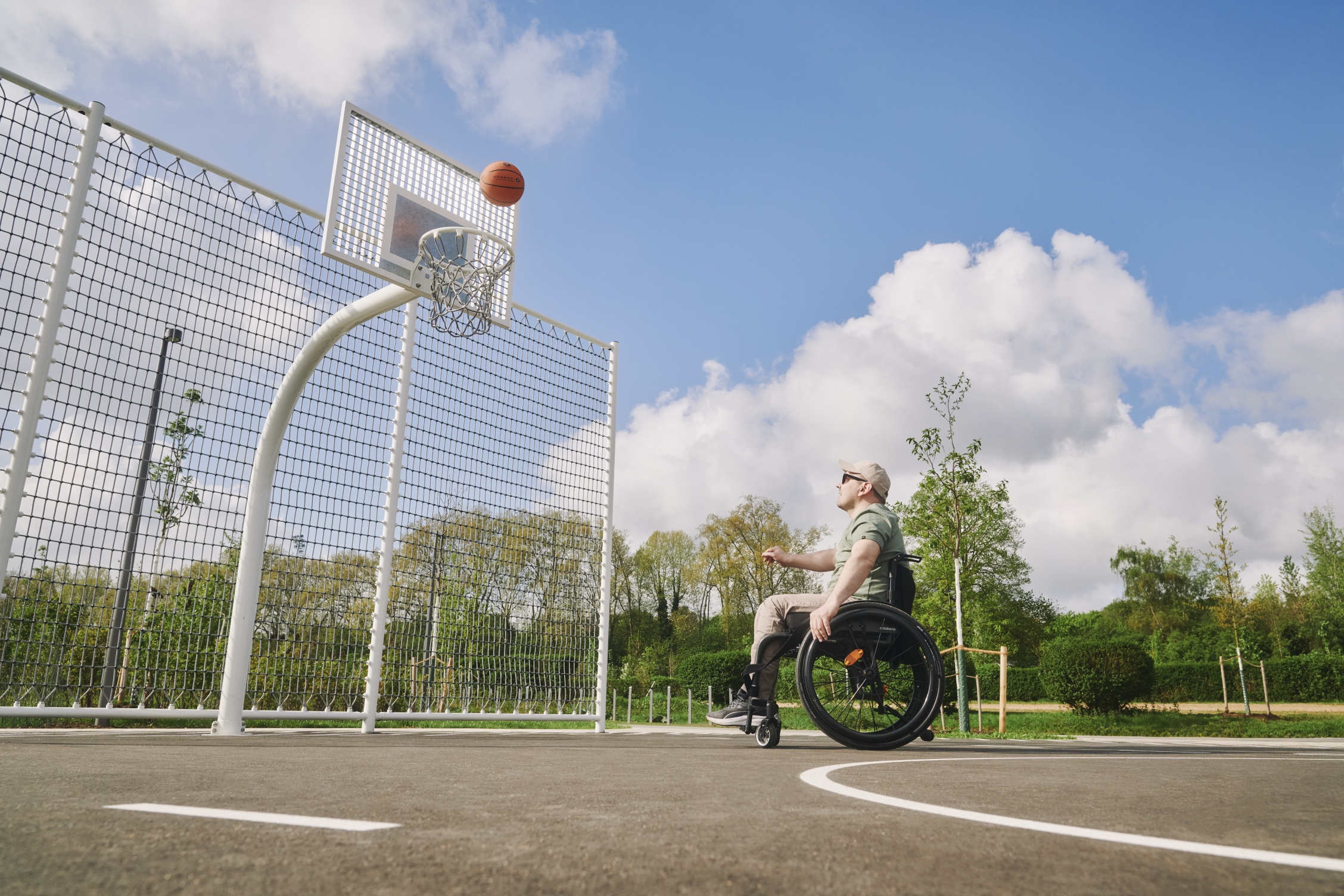 Ein Mann im Rollstuhl spielt auf einem Basketballplatz im Freien und wirft den Ball in Richtung Basketballkorb.
